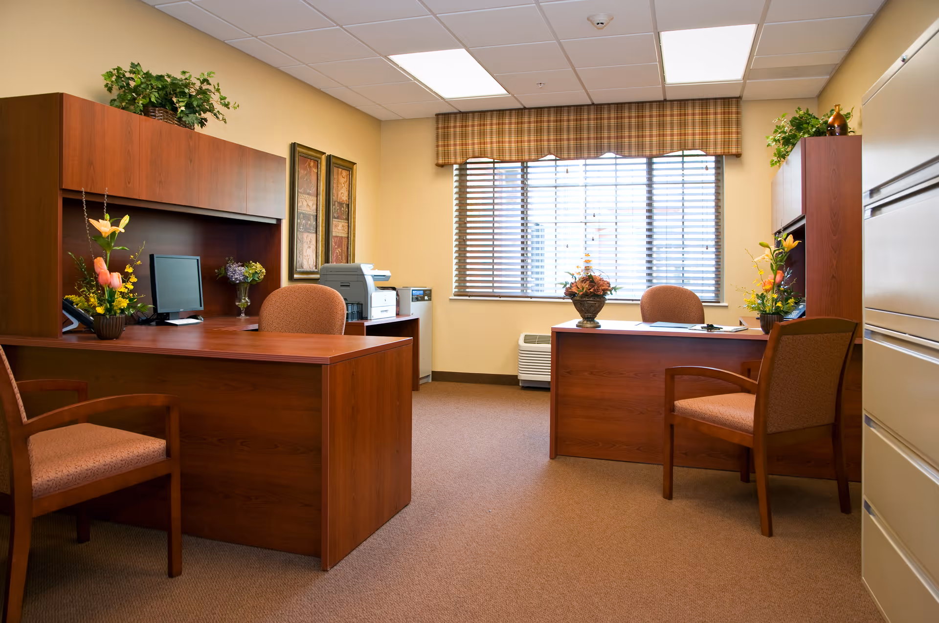 An office reception area with two wooden desks, upholstered chairs, computers, floral arrangements, and a window with blinds.