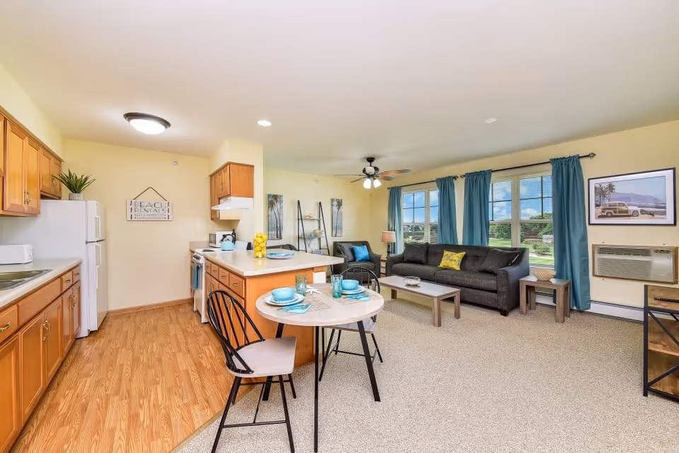 A bright and spacious open-concept living area and kitchen in a senior living facility. The kitchen features wooden cabinets, a white refrigerator, stove, and microwave, with a small island in the center. Adjacent to the kitchen is a small round dining table set with turquoise dishes and two chairs. The living room area has a dark gray sofa with colorful pillows, a coffee table, an armchair, and large windows with blue curtains letting in natural light. The walls are painted light yellow, and there is a ceiling fan with lights above the living room.