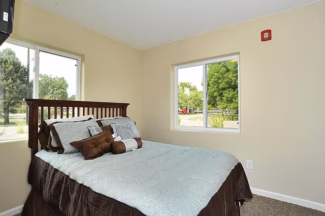 Bedroom with a neatly made bed against a wooden headboard and two windows showing trees outside.