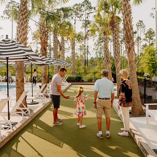 A family of four, including a young girl, a man, an older man, and an older woman, are playing bocce ball on a green court outdoors surrounded by palm trees. There are striped umbrellas and lounge chairs nearby, with a pool visible on the left side. The setting is sunny with a natural backdrop of trees and a black fence enclosing the area.
