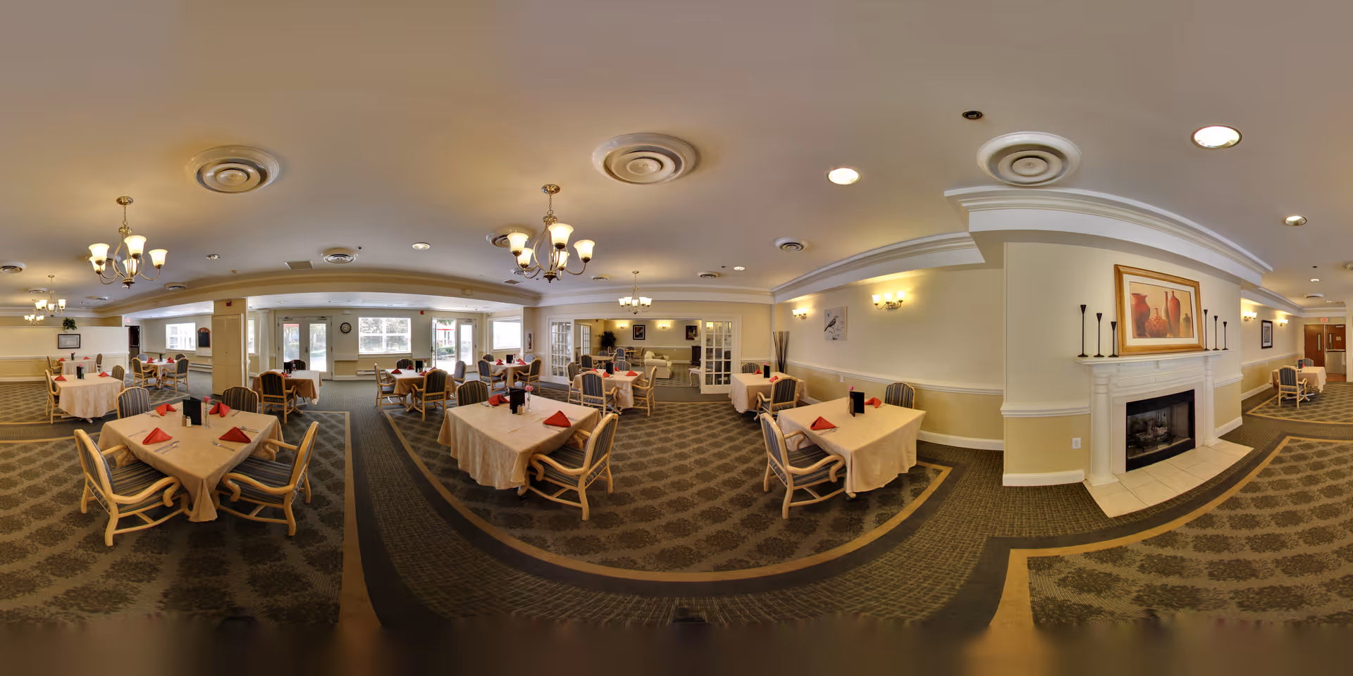 A spacious dining room in a senior living facility with multiple tables covered with beige tablecloths and set with red napkins. The room features patterned carpet, several chandeliers, a fireplace with a framed painting above it, and large windows letting in natural light. Chairs are arranged around the tables, and the overall ambiance is warm and inviting.