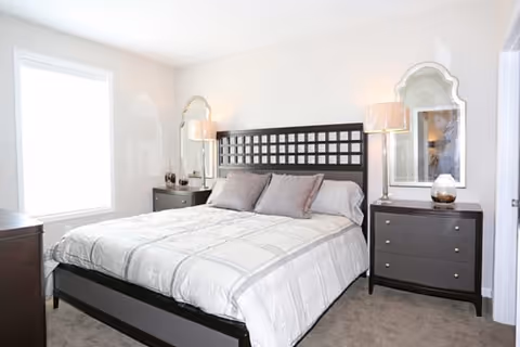 A bright bedroom with a large bed featuring a dark wooden headboard and light-colored bedding. There are two matching dark wooden nightstands on either side of the bed, each with a lamp and decorative items. Two ornate mirrors hang on the wall above the nightstands. A large window on the left side allows natural light to fill the room.