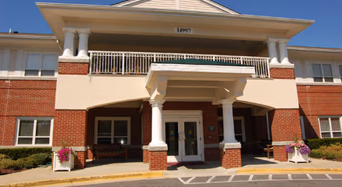 Brick two-story building entrance with white columns, a covered portico and balcony under a clear blue sky.