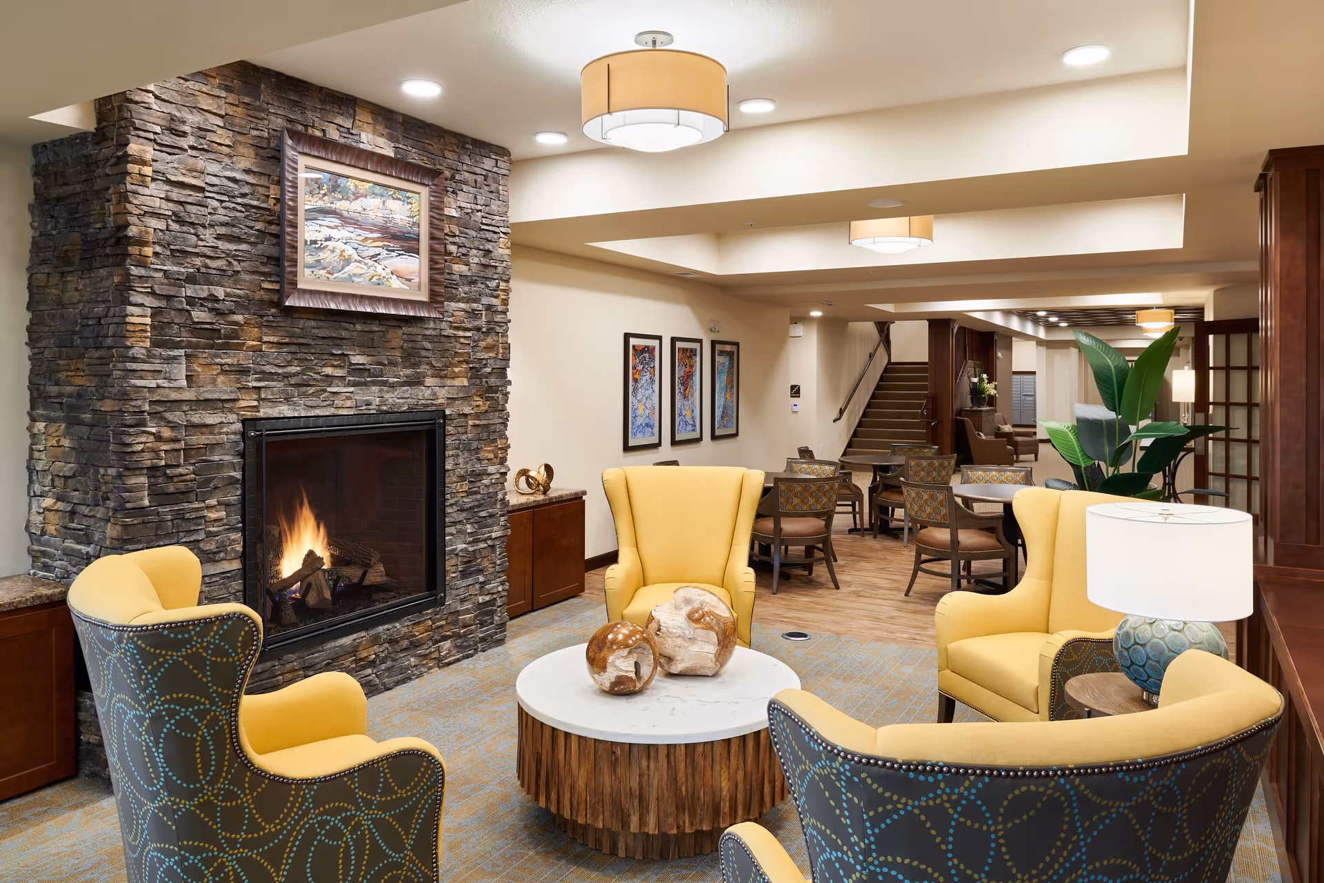 Cozy senior living common room with a stone fireplace, yellow armchairs around a round coffee table, and dining tables in the background.