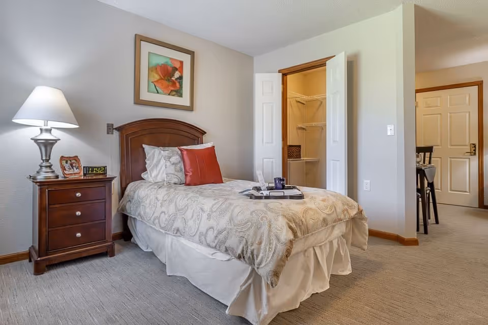 A neatly made single bed with patterned bedding and two pillows, one white and one red, in a bedroom. Next to the bed is a wooden nightstand with a silver lamp, a digital clock, and a framed photo. The room has light-colored walls and carpeted floor. An open closet with wire shelving is visible, as well as a doorway leading to another room with a dining table and chairs.