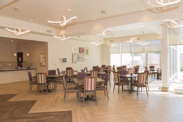 A spacious dining room with multiple tables and chairs arranged neatly. The chairs have striped upholstery in shades of purple and beige. Modern circular light fixtures hang from the ceiling. Large windows on the right side allow natural light to fill the room. The floor has a wood pattern with different shades creating a geometric design. The back wall has some framed artwork and a counter area with pendant lights above it.