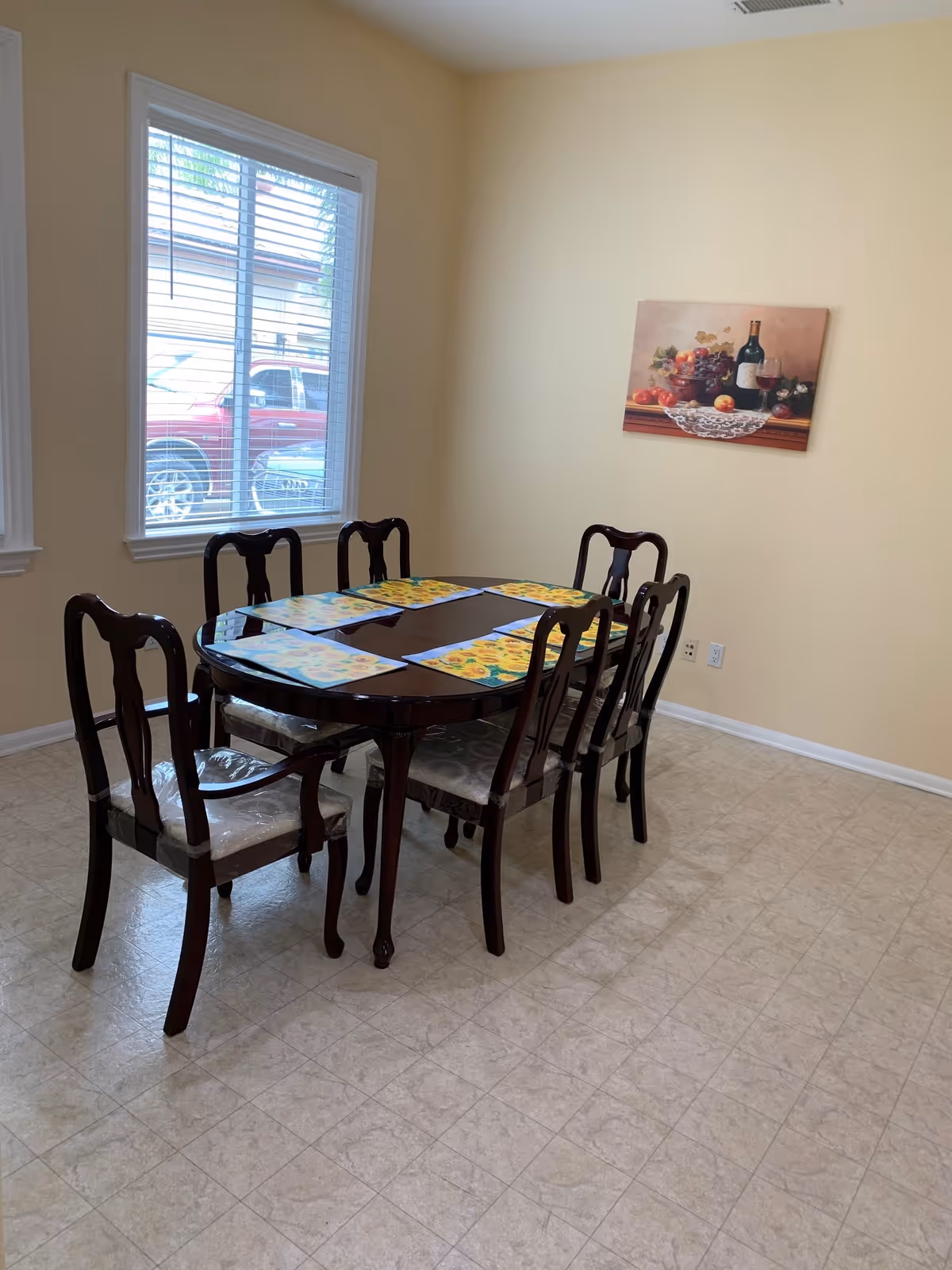 Sunlit dining room with a dark wooden table and six chairs set with colorful placemats, a window, and a fruit painting on a pale yellow wall.