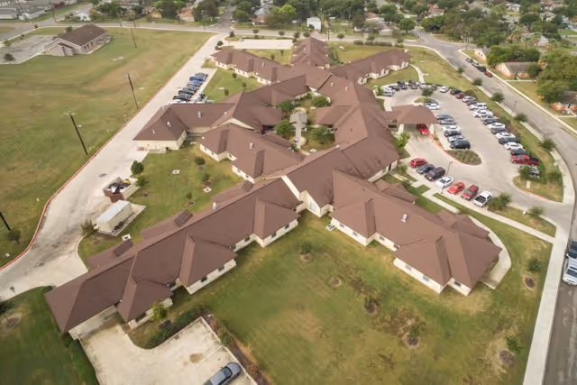 Aerial view of a large, single-story healthcare and rehabilitation facility with multiple wings and a brown roof, surrounded by parking lots, green lawns, and nearby residential areas.
