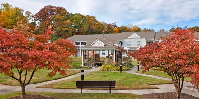 Outdoor view of a senior living facility named Rollingcrest Commons featuring a central gazebo surrounded by walkways, green lawns, and trees with vibrant red autumn foliage. A wooden bench is positioned in the foreground facing the gazebo, with a two-story building in the background under a partly cloudy sky.