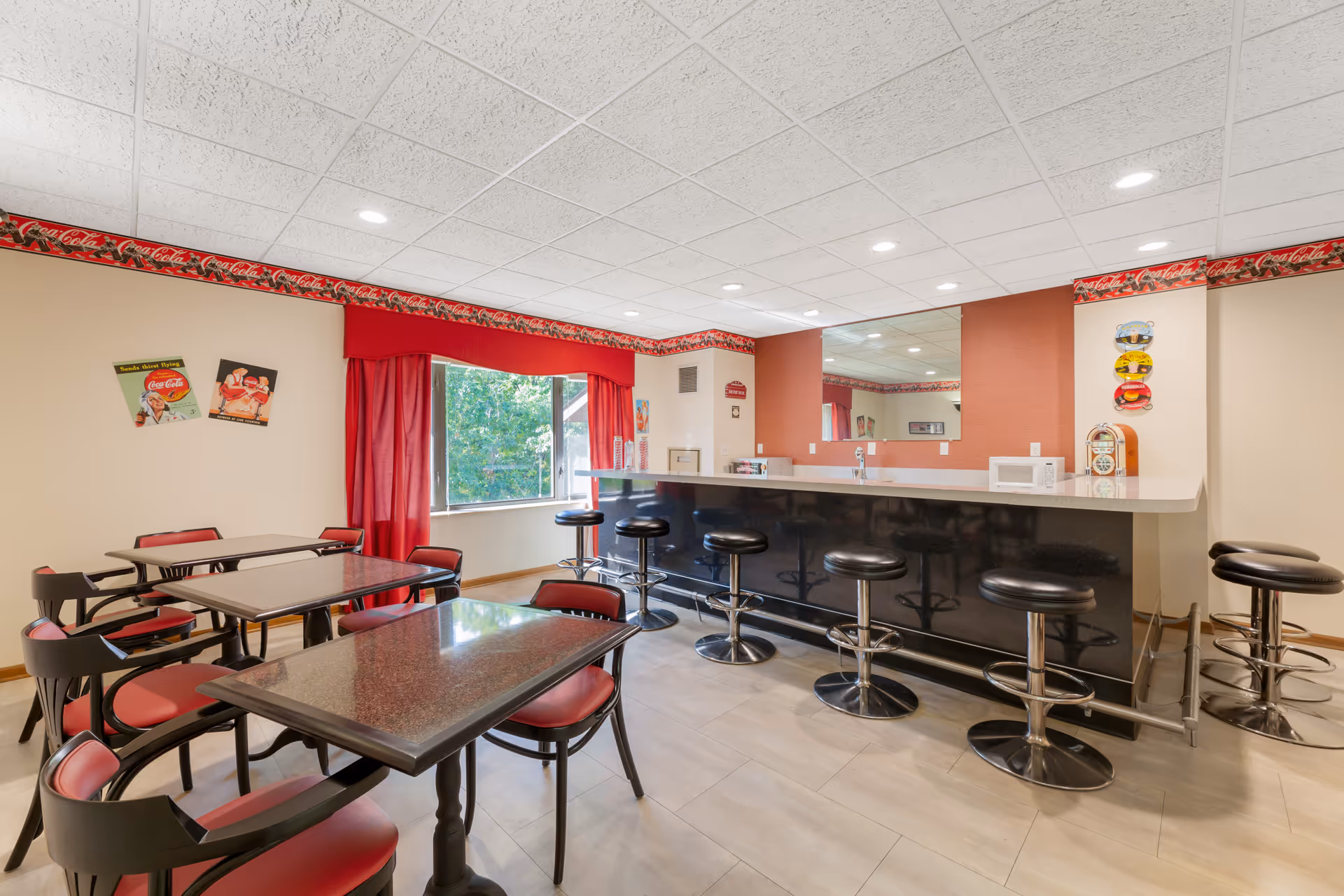 A retro-themed dining area with red and black chairs around tables and a bar counter with black bar stools. The room features Coca-Cola decorations including a border near the ceiling and vintage posters on the wall. There is a large window with red curtains letting in natural light, and a mirror behind the bar counter reflecting the room.