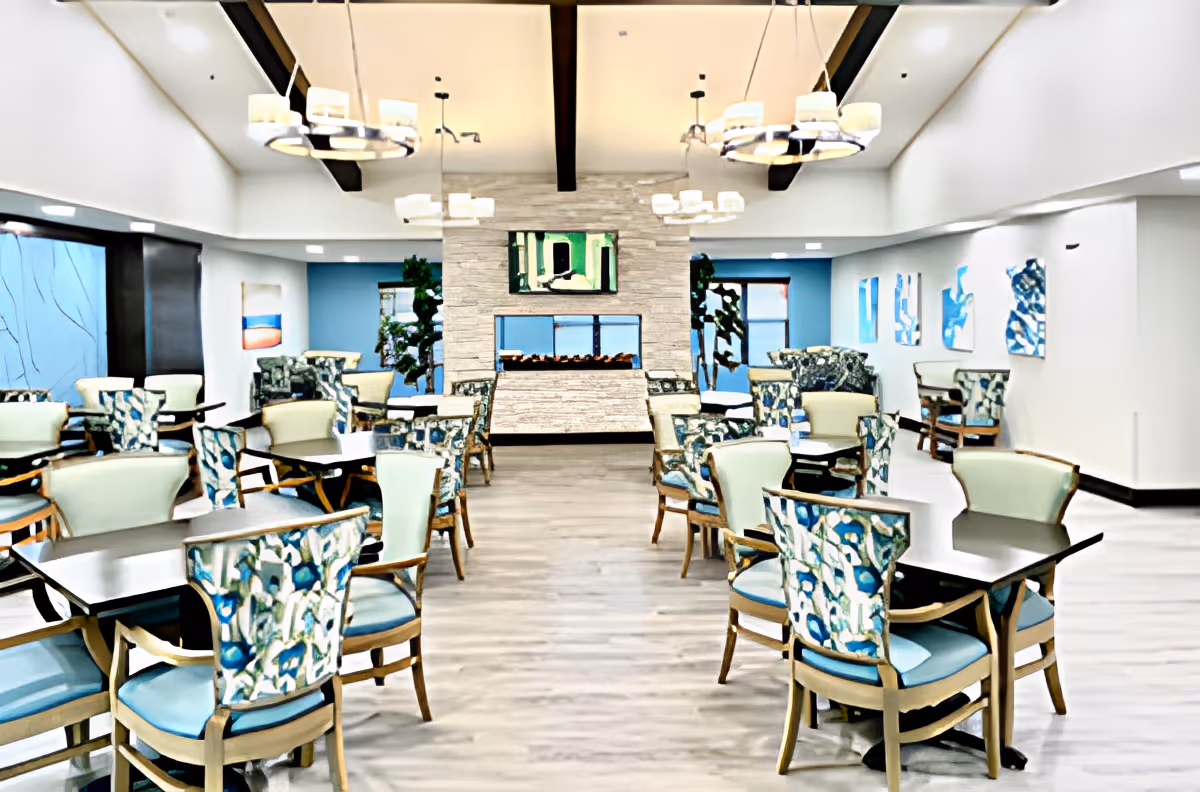 Bright, open dining area with multiple tables and patterned chairs facing a central stone fireplace and hanging chandeliers.