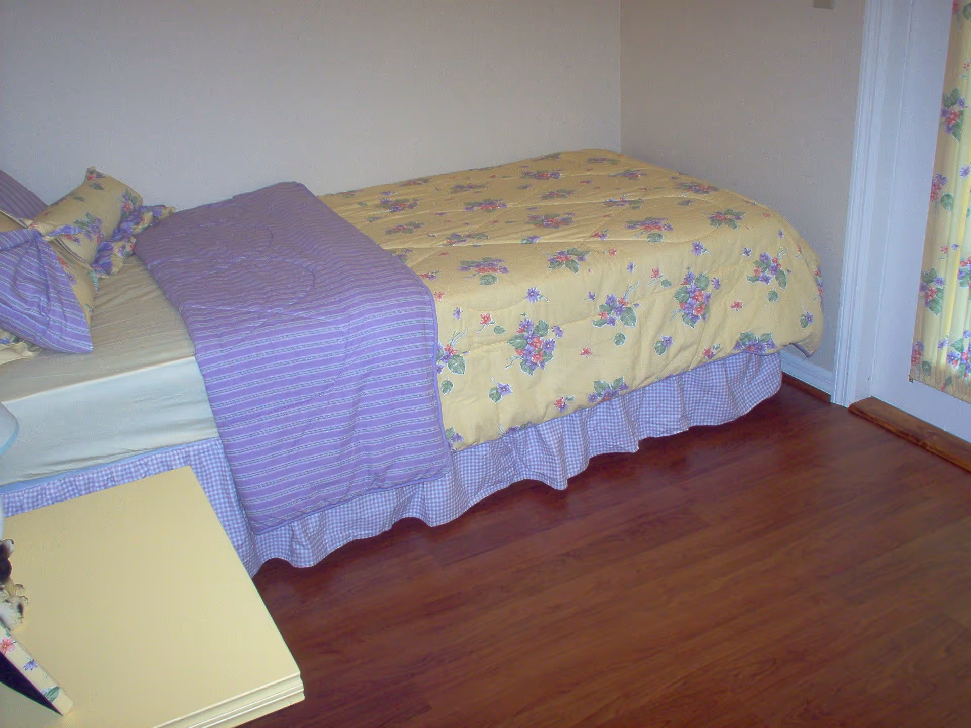 A bedroom with a bed covered in a yellow floral comforter and purple striped blanket, matching purple and yellow floral pillows, a yellow nightstand, and wood flooring. A door with a floral curtain is visible on the right side.