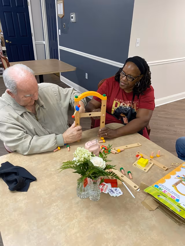 An elderly man and a woman sitting at a table in a room with blue and white walls. They are engaged in assembling a wooden toy or puzzle with colorful pegs. On the table, there is a small bouquet of white flowers in glass vases, some scattered toy pieces, and a box with colorful illustrations. The man is wearing a light beige jacket, and the woman is wearing glasses and a red shirt.