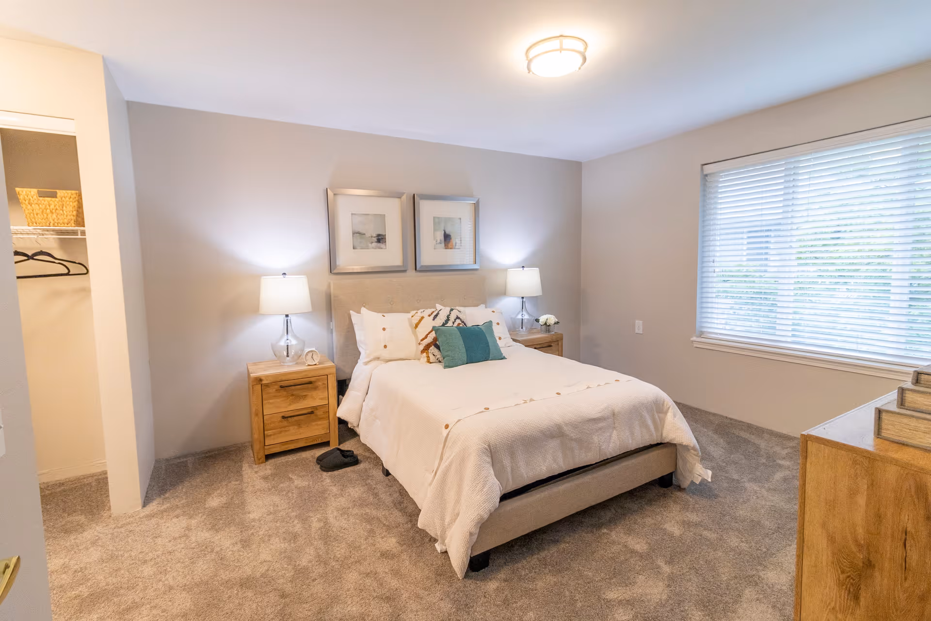 A cozy bedroom with a neatly made bed featuring white bedding and decorative pillows. There are two wooden nightstands on either side of the bed, each with a lamp. Two framed pictures hang above the bed. A window with blinds is on the right wall, and an open closet with hangers and a basket is visible on the left. The room has beige walls and carpeted flooring.