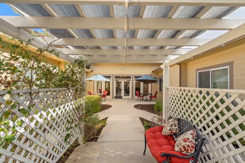 Covered outdoor walkway with lattice fencing on both sides, leading to a building entrance with glass doors. There are red cushioned chairs and tables with umbrellas along the walkway, and some greenery and plants around the area.