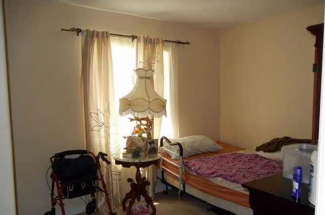 Small bedroom with a single bed, a bedside table holding a lamp and decorations, and a walker by the window.