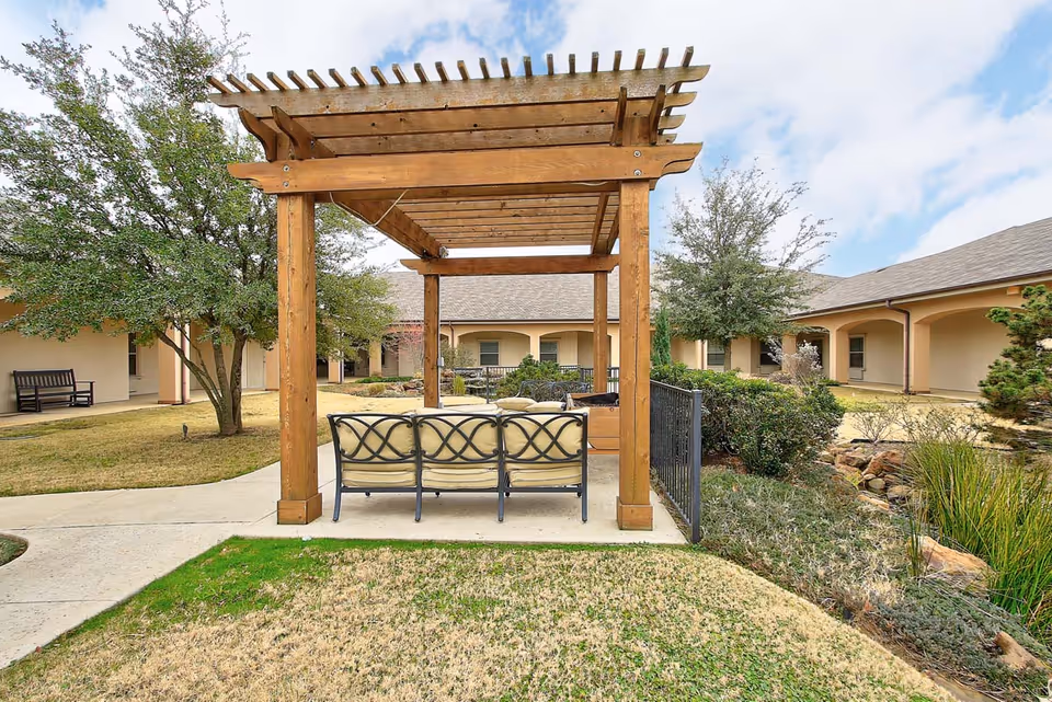 Outdoor seating area with cushioned metal chairs under a wooden pergola surrounded by grass, trees, and shrubs in a courtyard of a senior living facility.