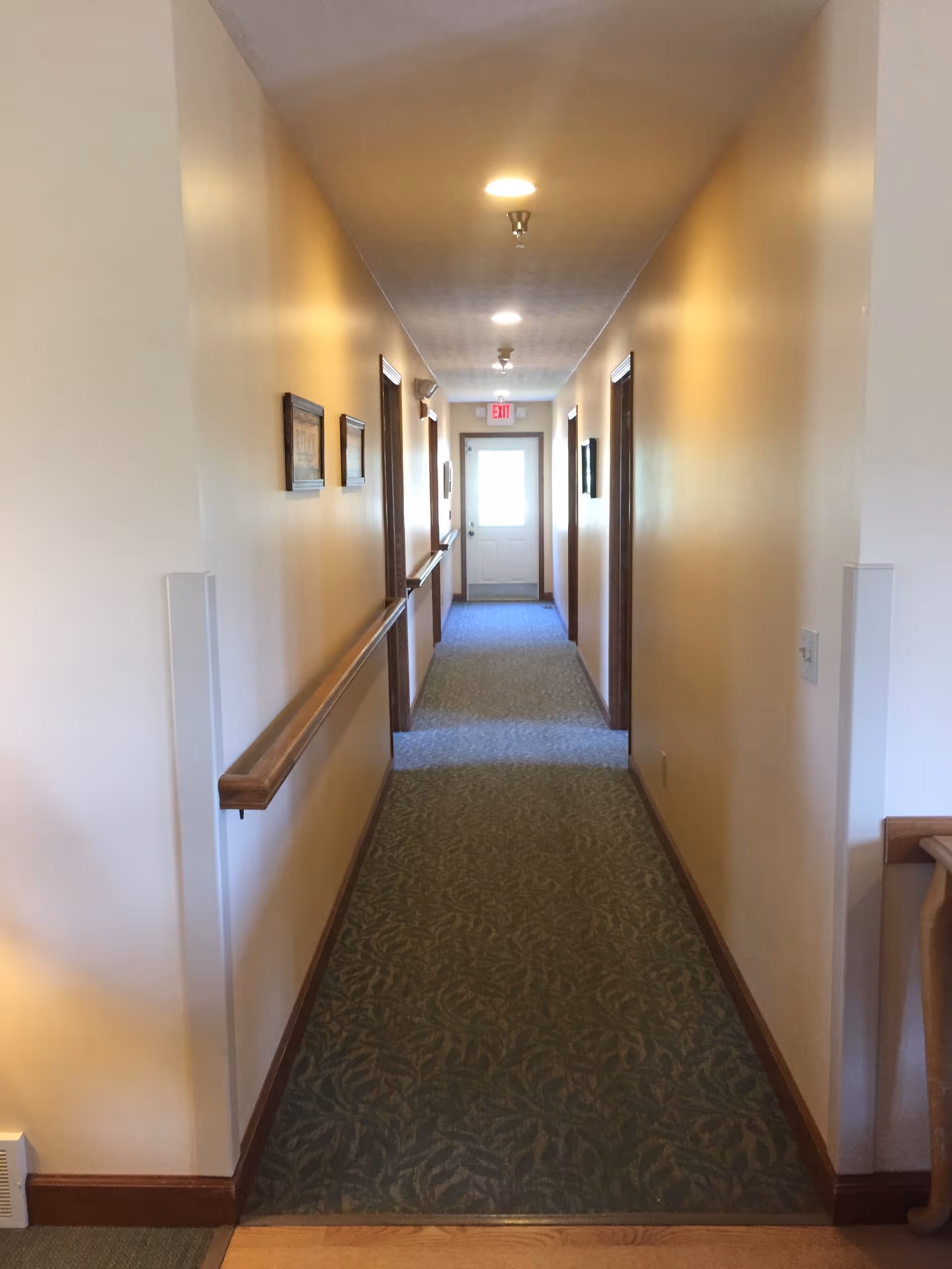 A long, narrow hallway in a senior living facility with beige walls, patterned carpet, wooden handrails on both sides, several doorways, framed pictures on the walls, and ceiling lights leading to a door with an exit sign above it.
