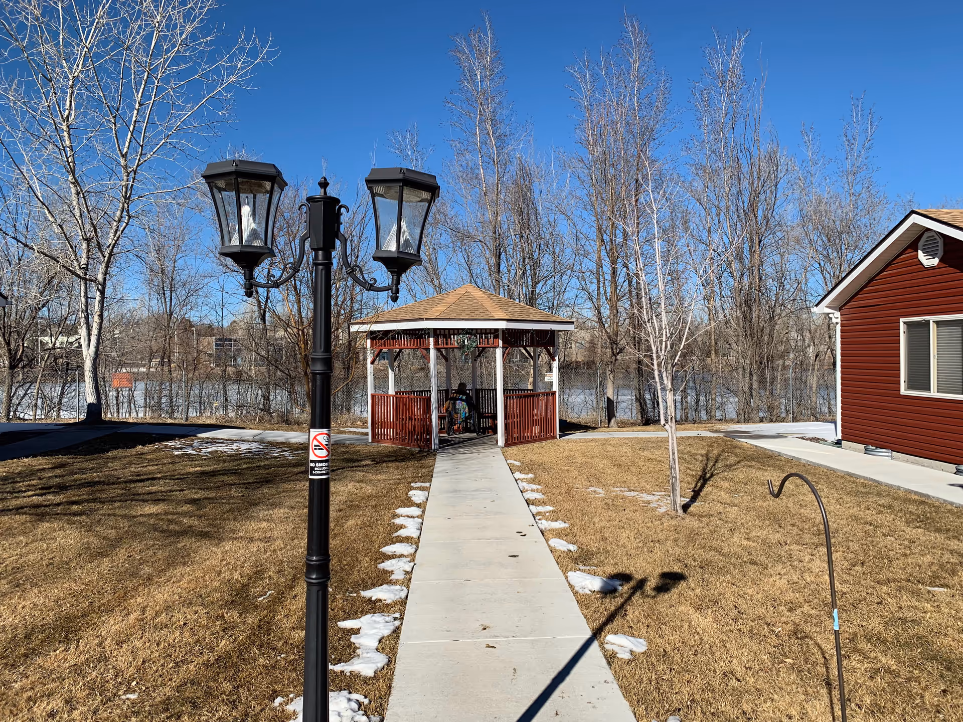 Outdoor scene at The Verandas Assisted Living showing a paved walkway leading to a wooden gazebo with a person sitting inside. The area is surrounded by leafless trees and patches of snow on the grass. A black lamp post with two lanterns and a no smoking sign is visible in the foreground, and a red building with white trim is on the right side under a clear blue sky.