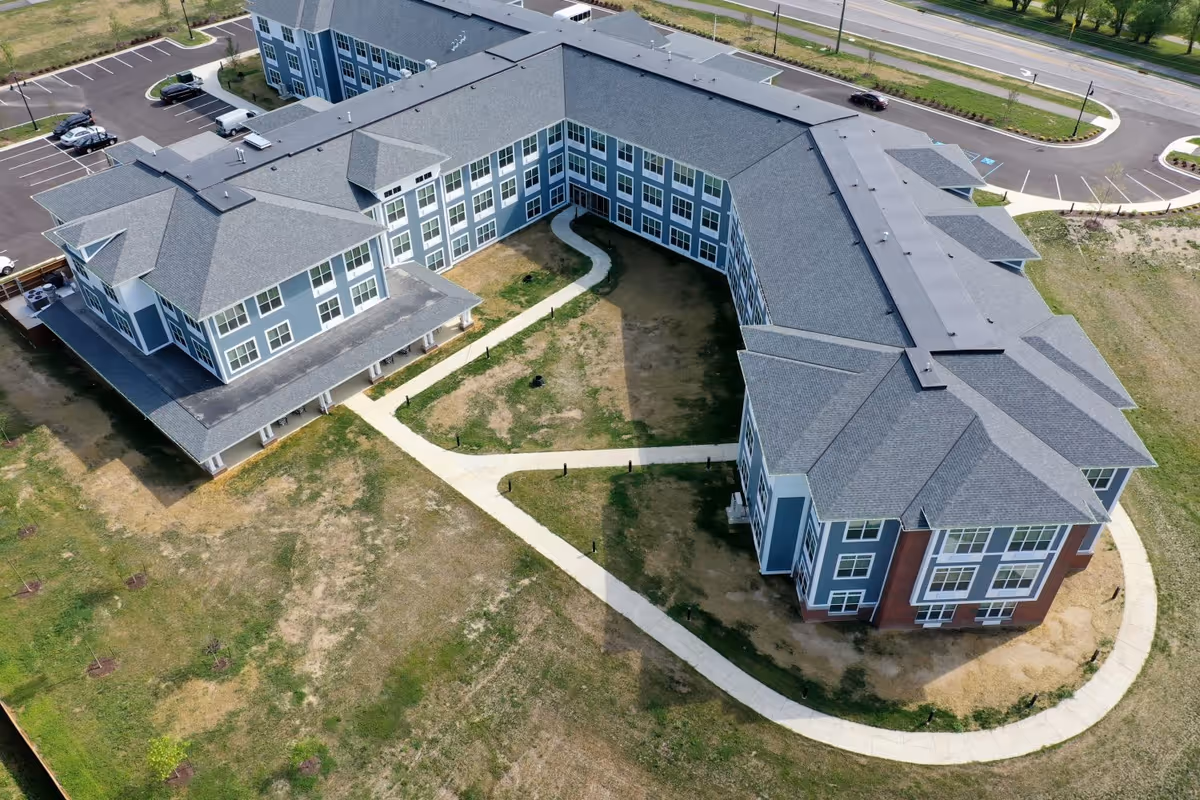 Aerial view of a large, modern senior living facility building with a U-shaped layout, surrounded by a paved walkway and grassy areas. The building has multiple windows and a gray roof, with a parking lot visible in the background.