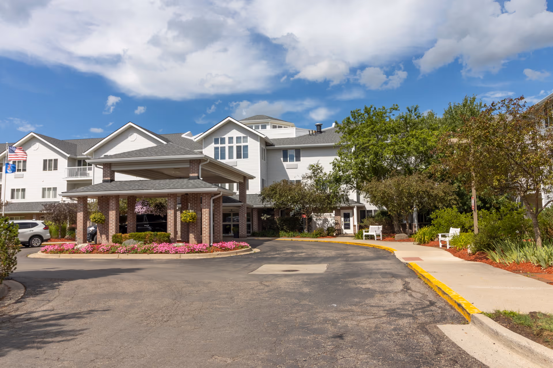 Exterior view of StoryPoint East Lansing senior living facility showing a multi-story building with white siding and a covered entrance supported by brick columns. There are landscaped flower beds with pink flowers, green shrubs, and trees around the entrance. A driveway curves in front of the building under a partly cloudy blue sky.