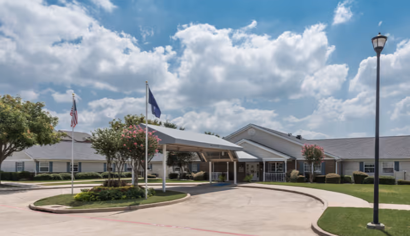 Exterior view of Arden Courts Of Richardson senior living facility showing a single-story building with a covered entrance, two flagpoles with flags, landscaped greenery, and a lamppost under a partly cloudy sky.