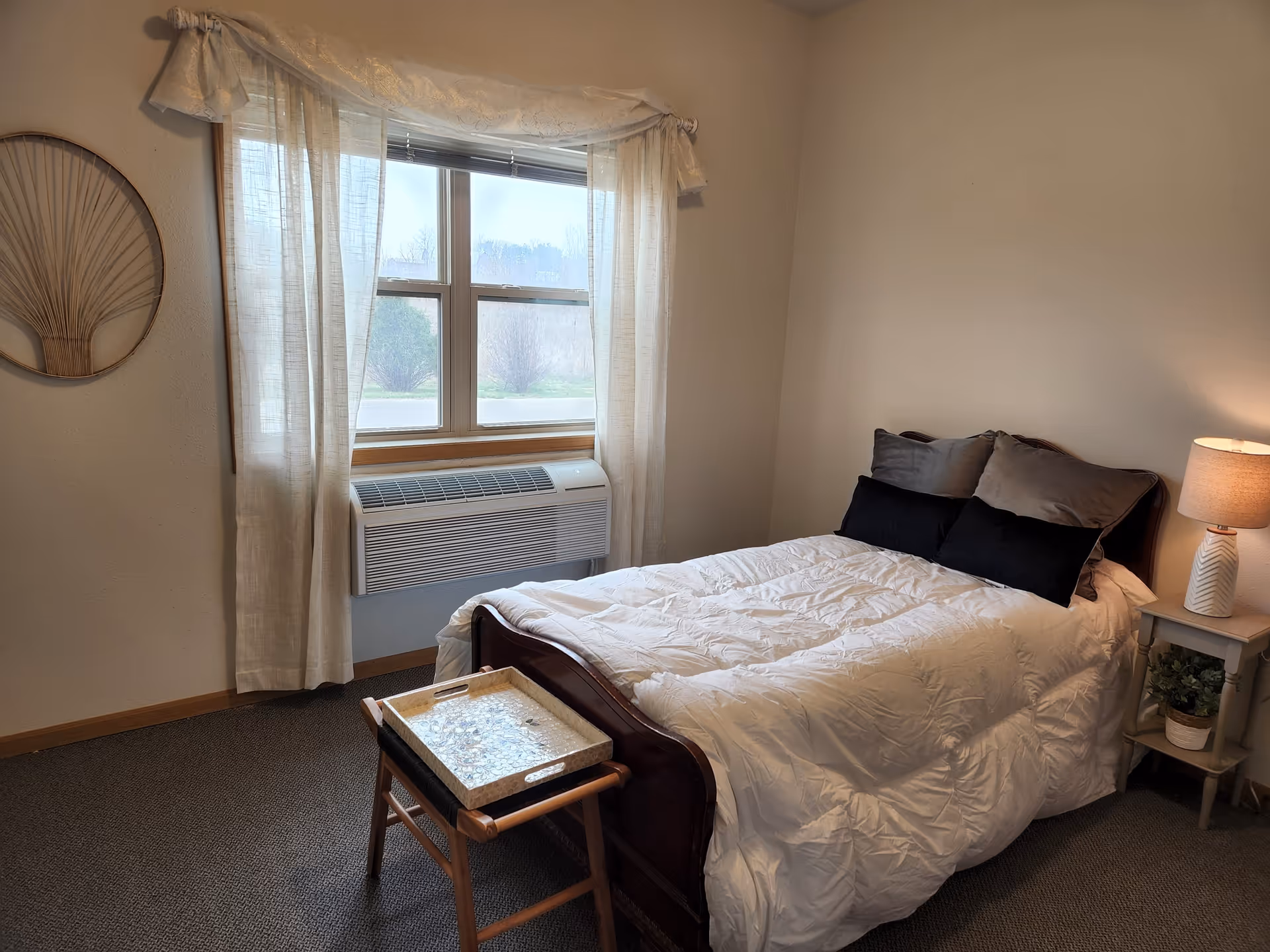 A cozy bedroom with a neatly made bed featuring white bedding and four pillows in black and gray. There is a small wooden tray table at the foot of the bed. A window with sheer white curtains lets in natural light, and below the window is a wall-mounted air conditioning unit. A nightstand with a lamp and a small plant is beside the bed. The walls are light-colored, and there is a decorative wall hanging on the left side.