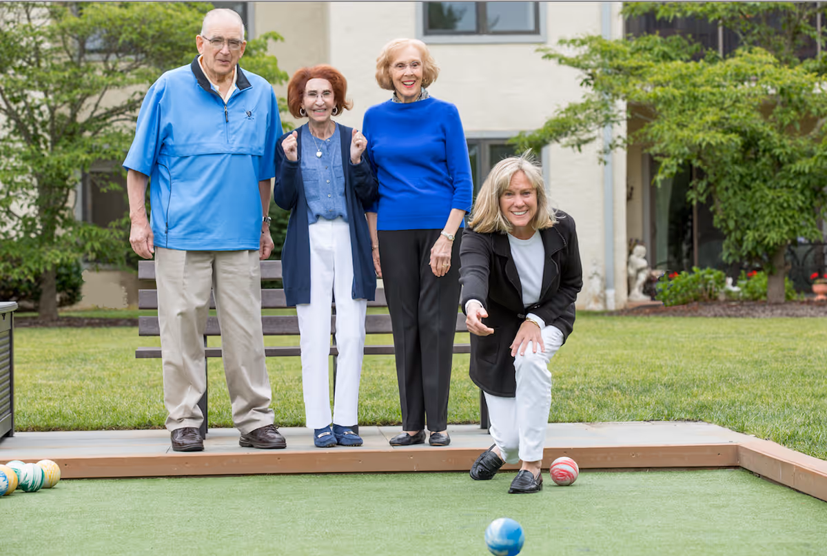 Four elderly adults enjoying a game of bocce ball outdoors on a green court. Three of them are standing and watching while one woman is crouched down, rolling a bocce ball. They are in a garden area with grass, trees, and a building in the background.