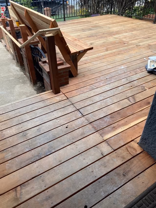 A wooden outdoor deck with built-in wooden benches along the edge. The deck has a railing and overlooks a grassy area with some plants and a brick structure in the background.