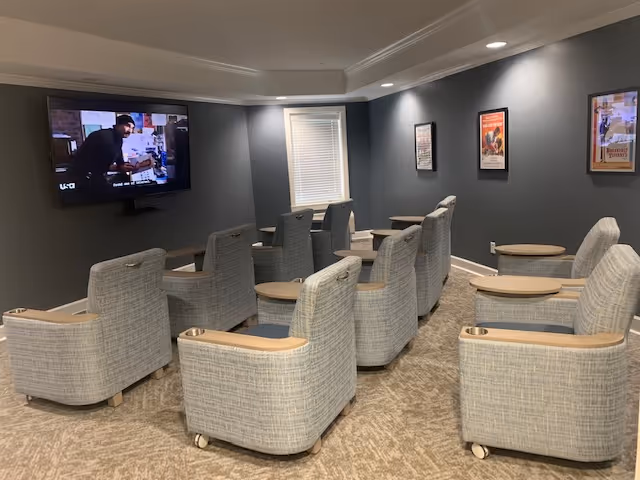 A small theater room with two rows of comfortable, light gray upholstered chairs with wooden armrests and cup holders facing a wall-mounted flat screen TV. The walls are painted dark gray and decorated with framed movie posters. There is a window with blinds on the back wall and recessed ceiling lights illuminating the room.