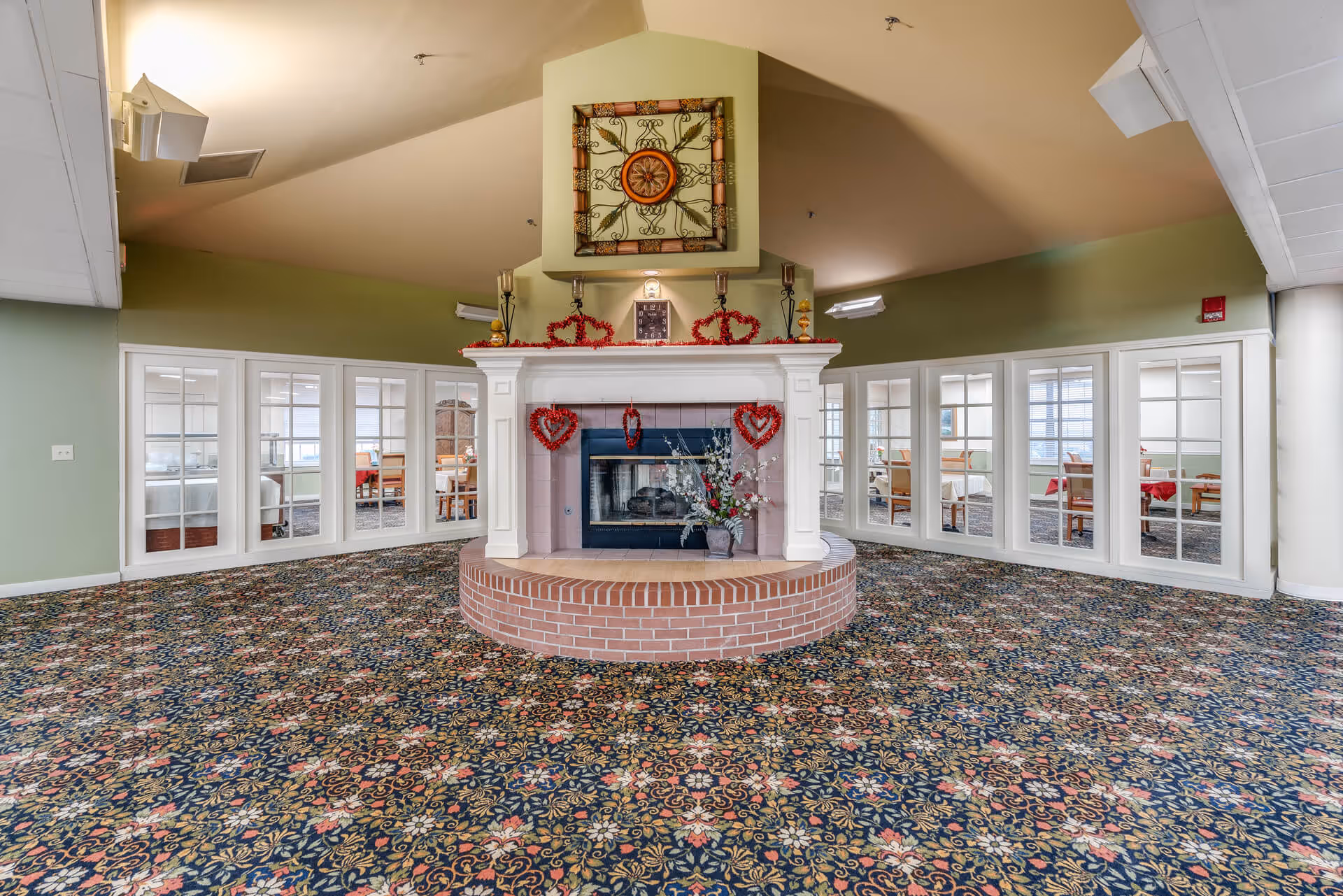 Interior view of a senior living facility common area with a central white fireplace decorated with red heart-shaped garlands and floral arrangements. The room has a patterned carpet and green walls with large windows and tables with chairs visible through glass panels on either side of the fireplace.