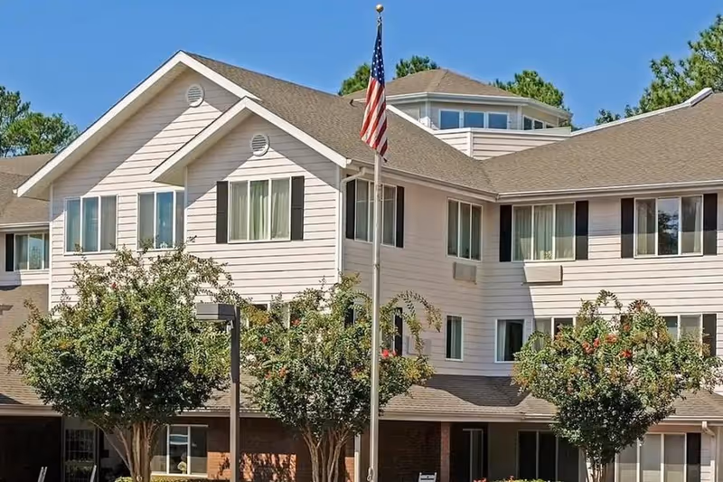 Sunlit multi-story building with white siding, an American flag on a flagpole, and trees in front.