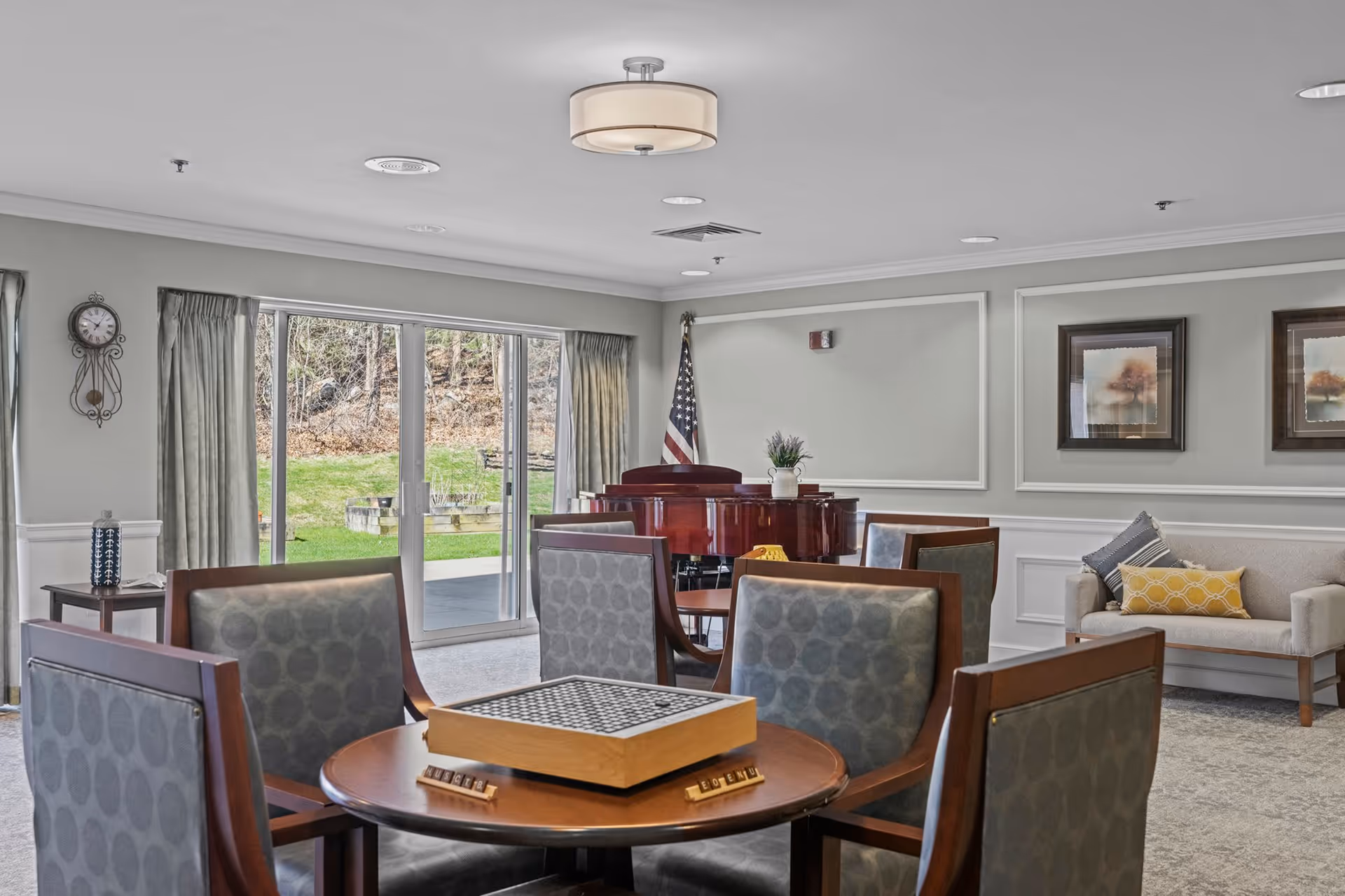A senior living facility common area with a round table and four chairs, a chess/checkers board on the table, a beige couch with decorative pillows, framed artwork on the wall, an American flag, a red piano, and large sliding glass doors showing an outdoor grassy area.