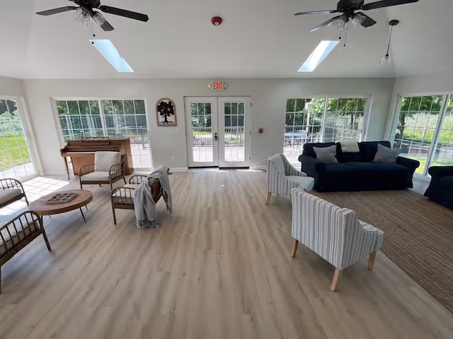 Bright communal lounge with sofas, chairs, a piano, large windows, and double doors leading outside.