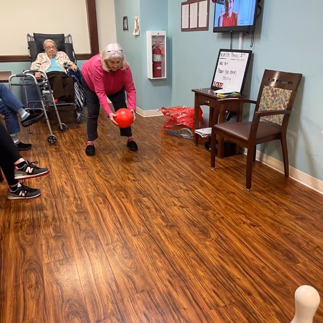 An elderly woman in a pink top is bending down to roll a red ball on a wooden floor in a room. Other elderly individuals are seated around the room, some in wheelchairs. There is a small table with a chair against the wall, a fire extinguisher mounted on the wall, and a TV screen displaying a person. A whiteboard with writing is also visible near the table.