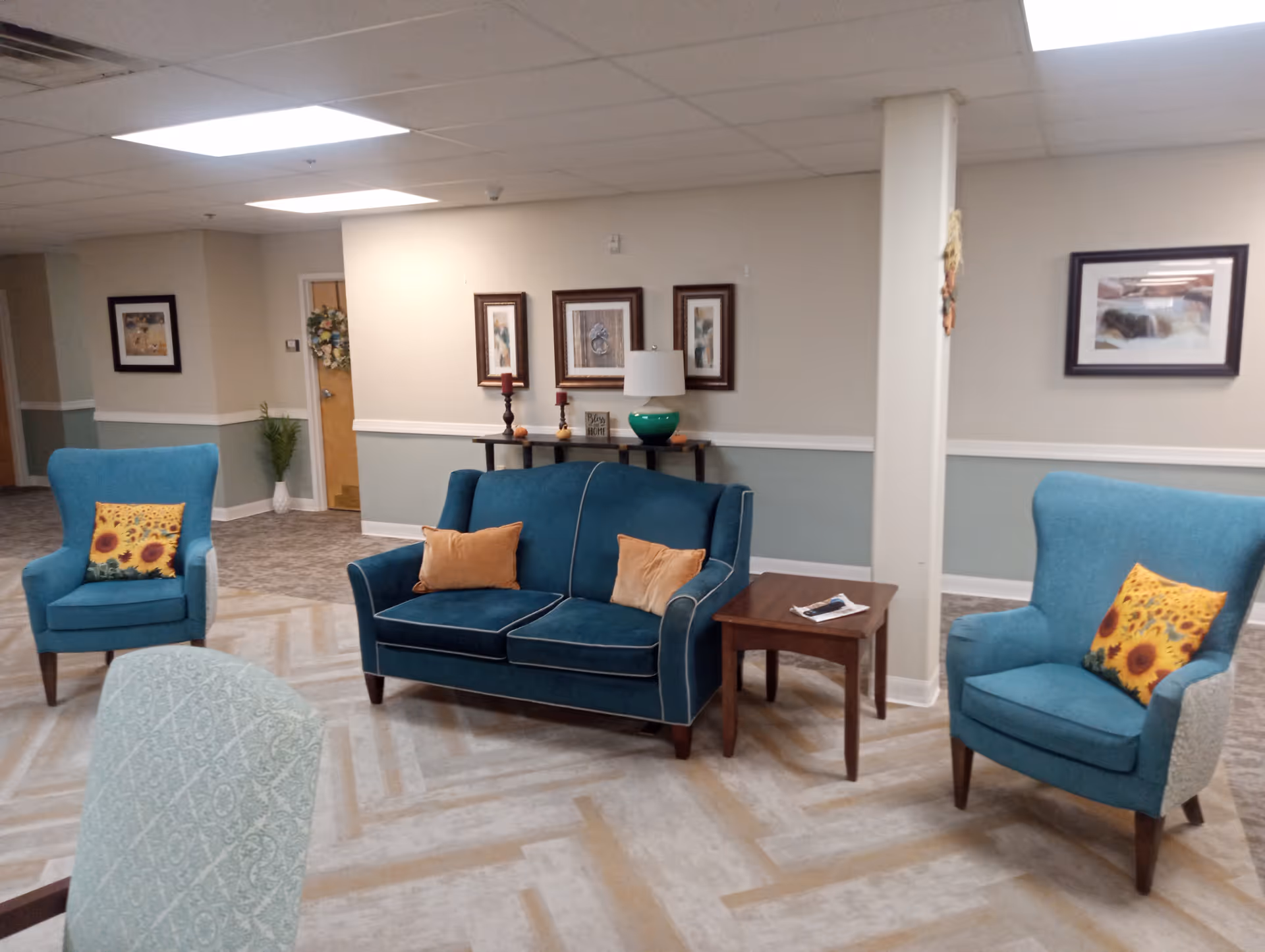 A cozy seating area in a senior living facility with a teal loveseat and two matching teal armchairs, each armchair has a sunflower-patterned pillow. There is a wooden side table with a remote control and a console table against the wall with decorative items including candles, a lamp, and framed artwork. The walls are painted beige with a light green wainscoting, and the floor has a patterned carpet.