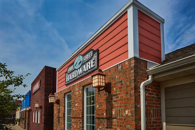 Exterior view of a brick building with a sign that reads 'Forest Street Hardware' under a blue sky with some clouds. The building features red siding on the upper portion and two lantern-style wall lights beside a window.