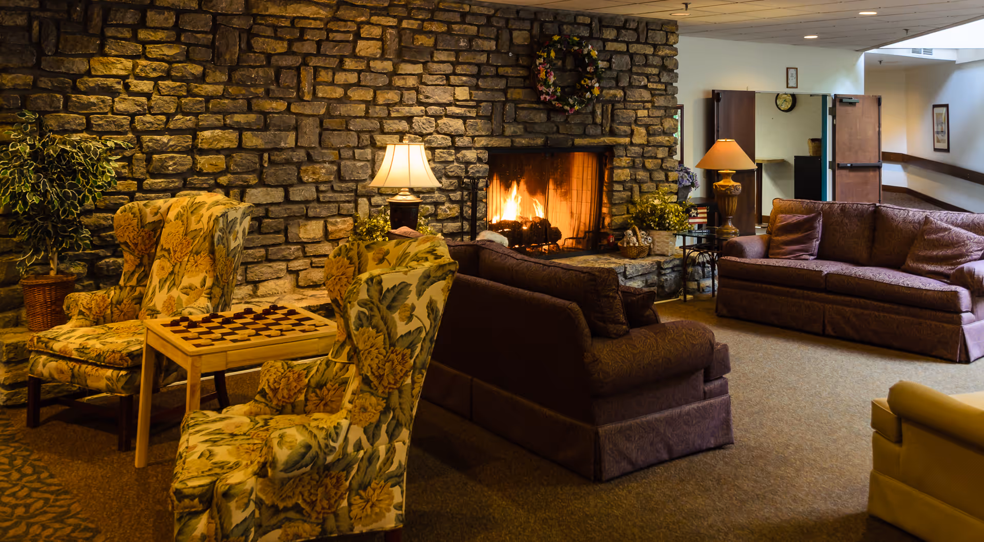 A cozy living room area with a stone fireplace burning brightly. The room features two floral-patterned armchairs with a wooden table between them that has a checkerboard on top. There are two brown sofas facing the fireplace, a couple of table lamps, and a potted plant. The walls are decorated with a wreath above the fireplace and framed pictures. The space has a warm and inviting atmosphere.