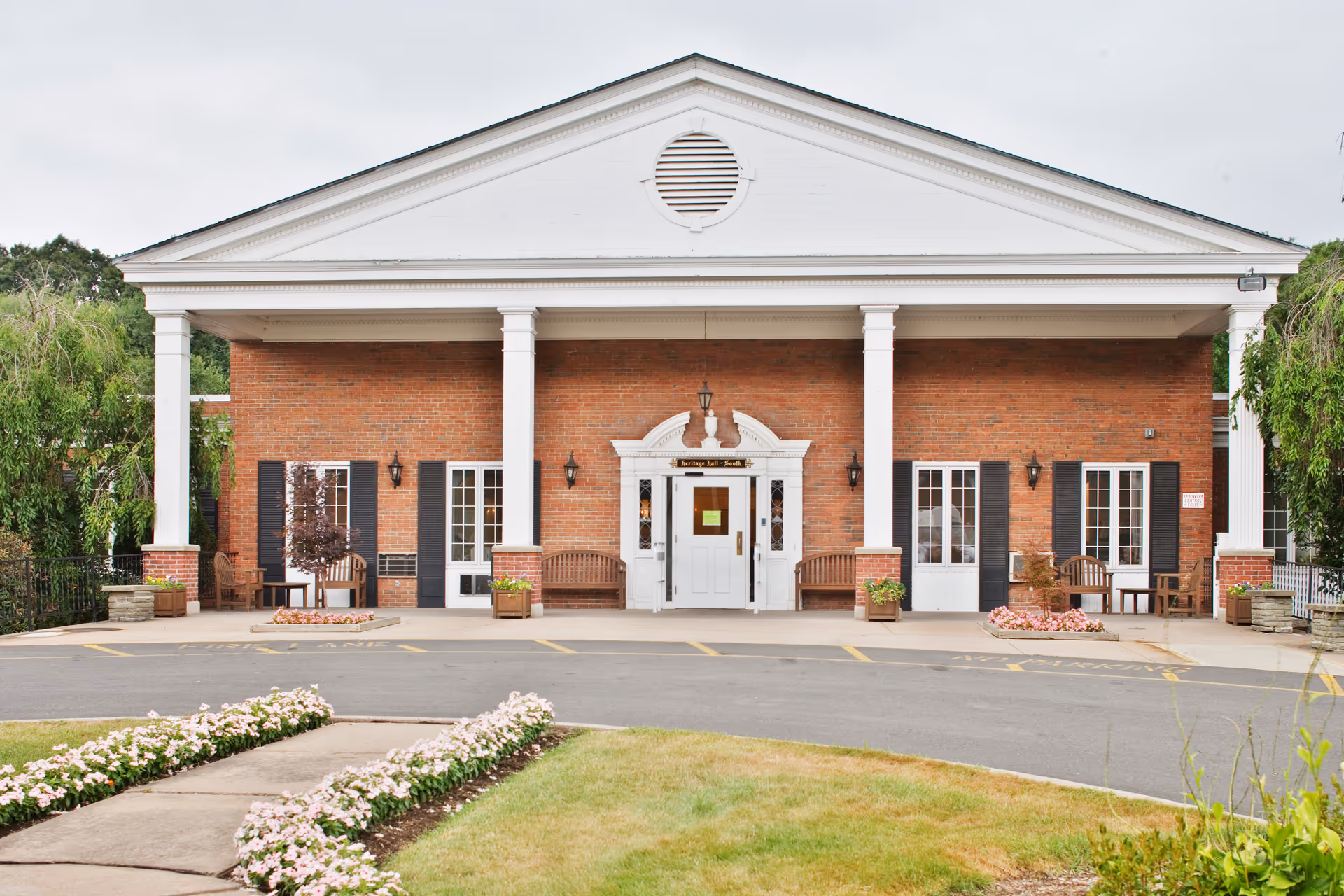 Front exterior view of a brick building with white columns and a triangular pediment. The entrance has a white door with sidelights and a decorative white frame. There are benches and potted plants on either side of the entrance, and flower beds line the walkway leading to the building.
