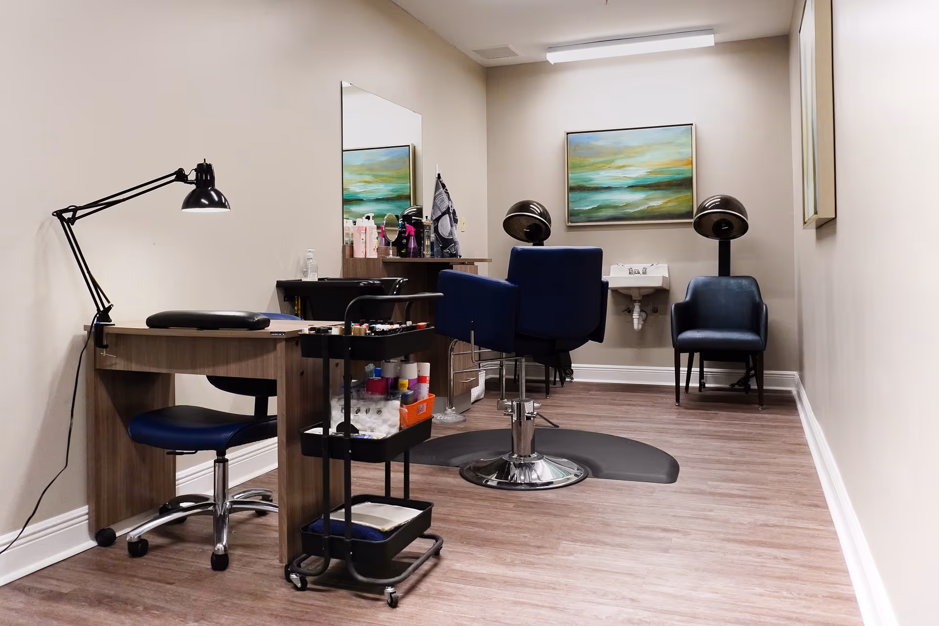 Interior view of a salon area in a senior living facility featuring a manicure table with a black adjustable lamp and a blue swivel chair. In the background, there is a hair styling station with a blue salon chair, a hair dryer hood, a small sink, and a blue armchair. The walls are beige with two framed paintings, and the floor has wood-like vinyl flooring.