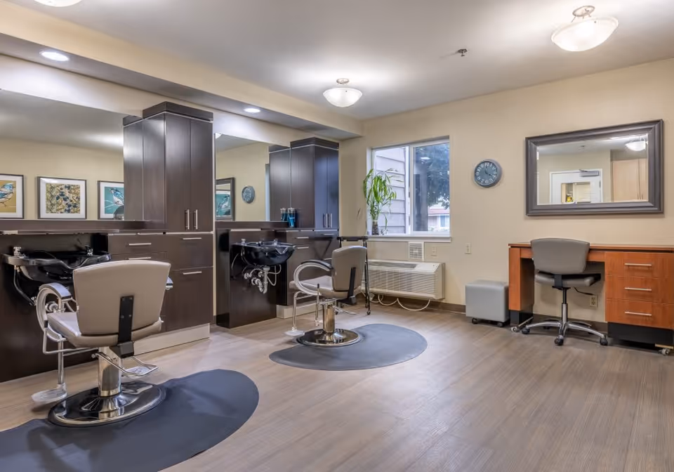 Interior view of a hair salon area in a senior living facility with two salon chairs in front of black wash basins and dark wood cabinetry. There is a large mirror on the wall behind the chairs, a window letting in natural light, a clock on the wall, and a desk with a chair and a mirror on the right side of the room.