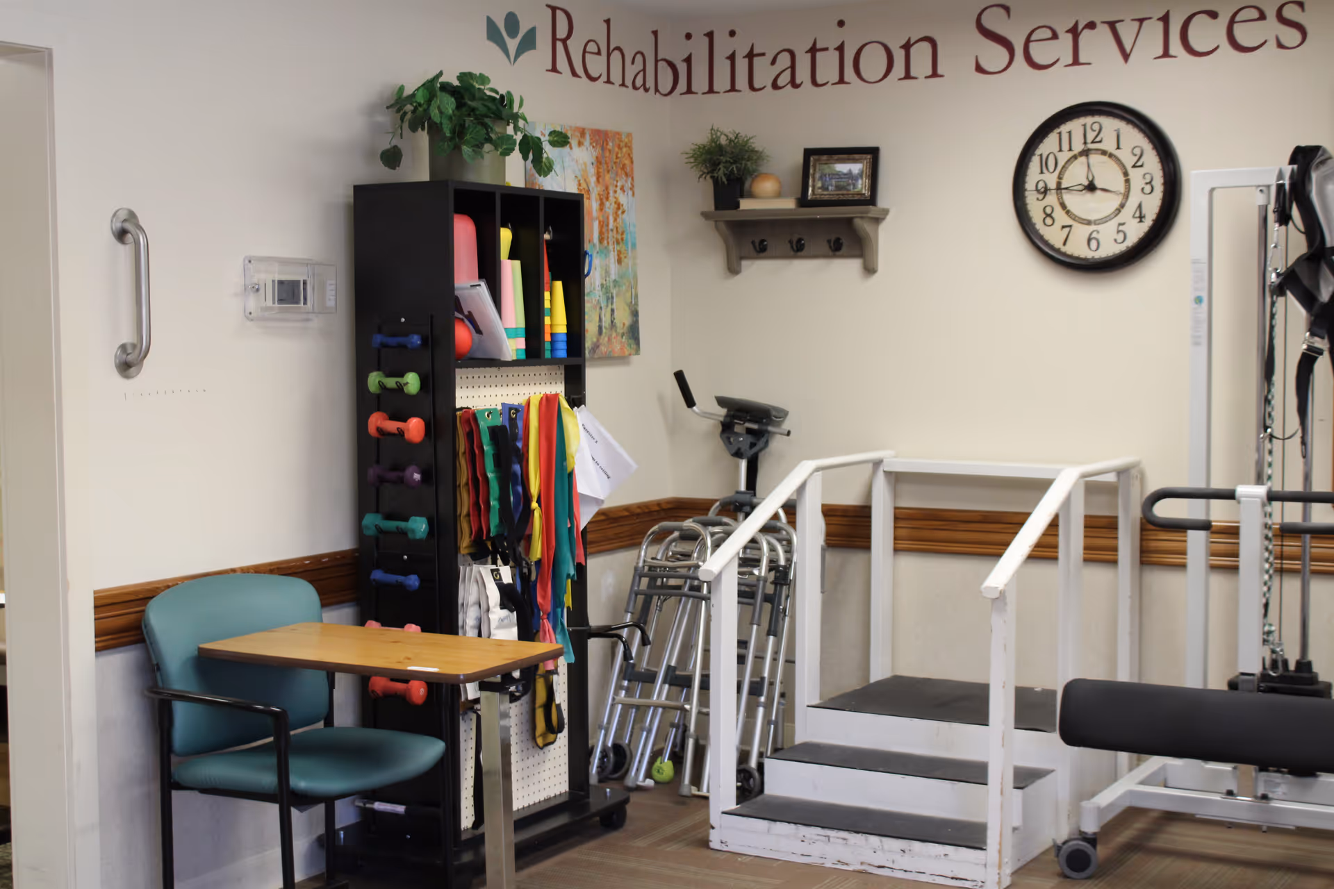 A rehabilitation services room with exercise equipment including a small set of stairs with handrails, walkers, resistance bands, dumbbells, and a stationary bike. There is a teal chair with a wooden table attached, a wall clock, a shelf with hooks, and a plant on top of a black storage unit. The words 'Rehabilitation Services' are partially visible on the wall.