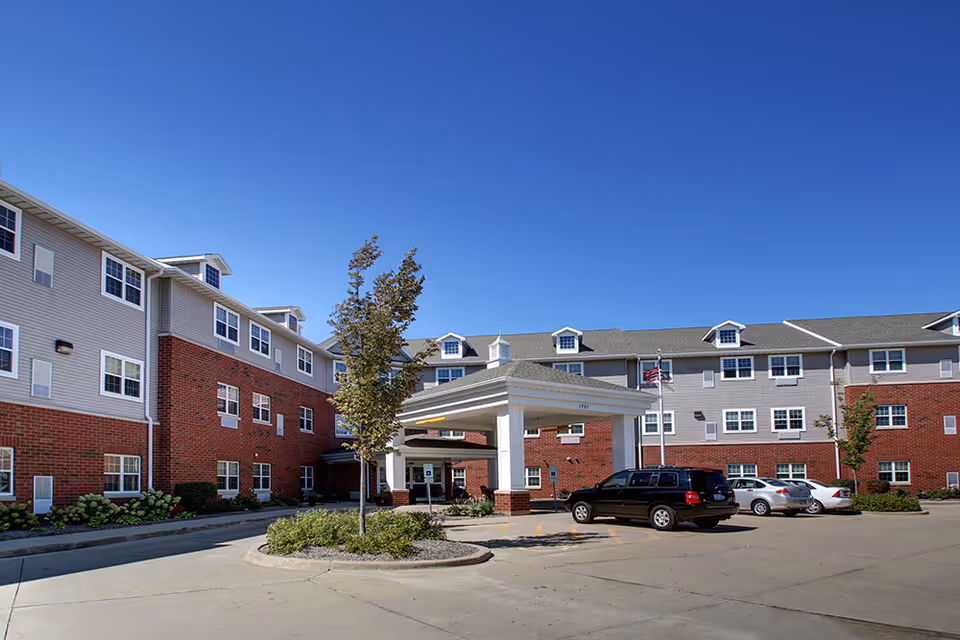 Exterior view of a senior living facility building with a covered entrance, several parked cars, and a clear blue sky.