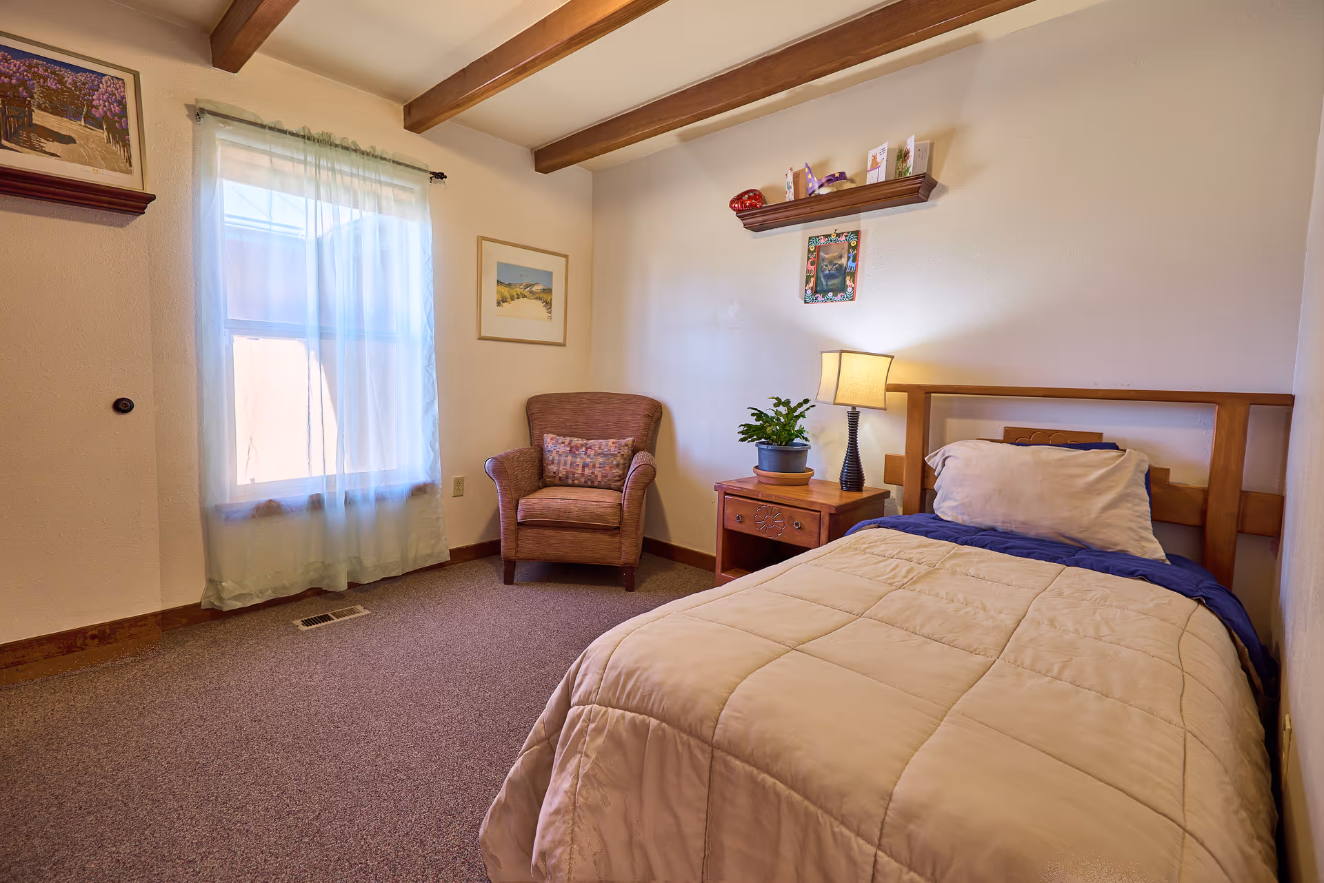 A cozy bedroom with a single bed covered in a beige comforter and a blue blanket, a wooden headboard, a wooden nightstand with a lamp and a potted plant, a comfortable armchair with a decorative pillow, two framed pictures on the walls, a window with sheer curtains, and wooden ceiling beams.