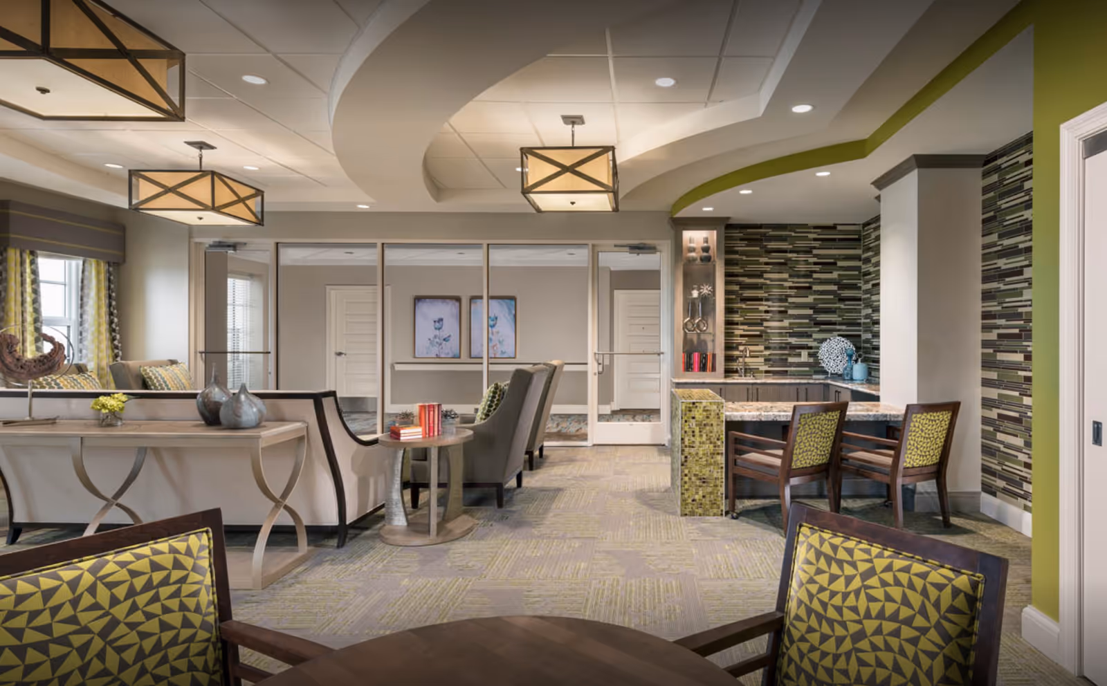 A modern, open senior living common area with seating, tables, pendant lights, and a tiled accent wall.