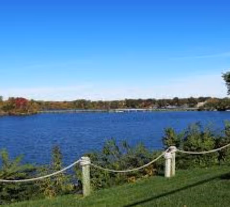 A scenic view of a large blue lake with green bushes and grass in the foreground, a rope fence with wooden posts along the edge, and a clear blue sky above.