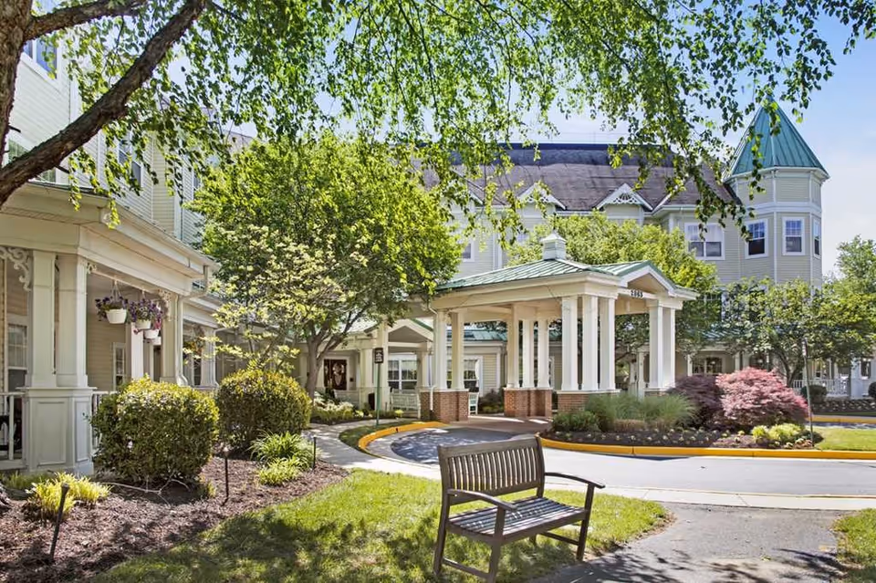 Outdoor view of Sunrise at Hunter Mill facility showing a circular driveway with a covered entrance supported by white columns. The building is light-colored with multiple windows and a green roof turret. There are trees, shrubs, and a wooden bench in the landscaped garden area under a clear blue sky.