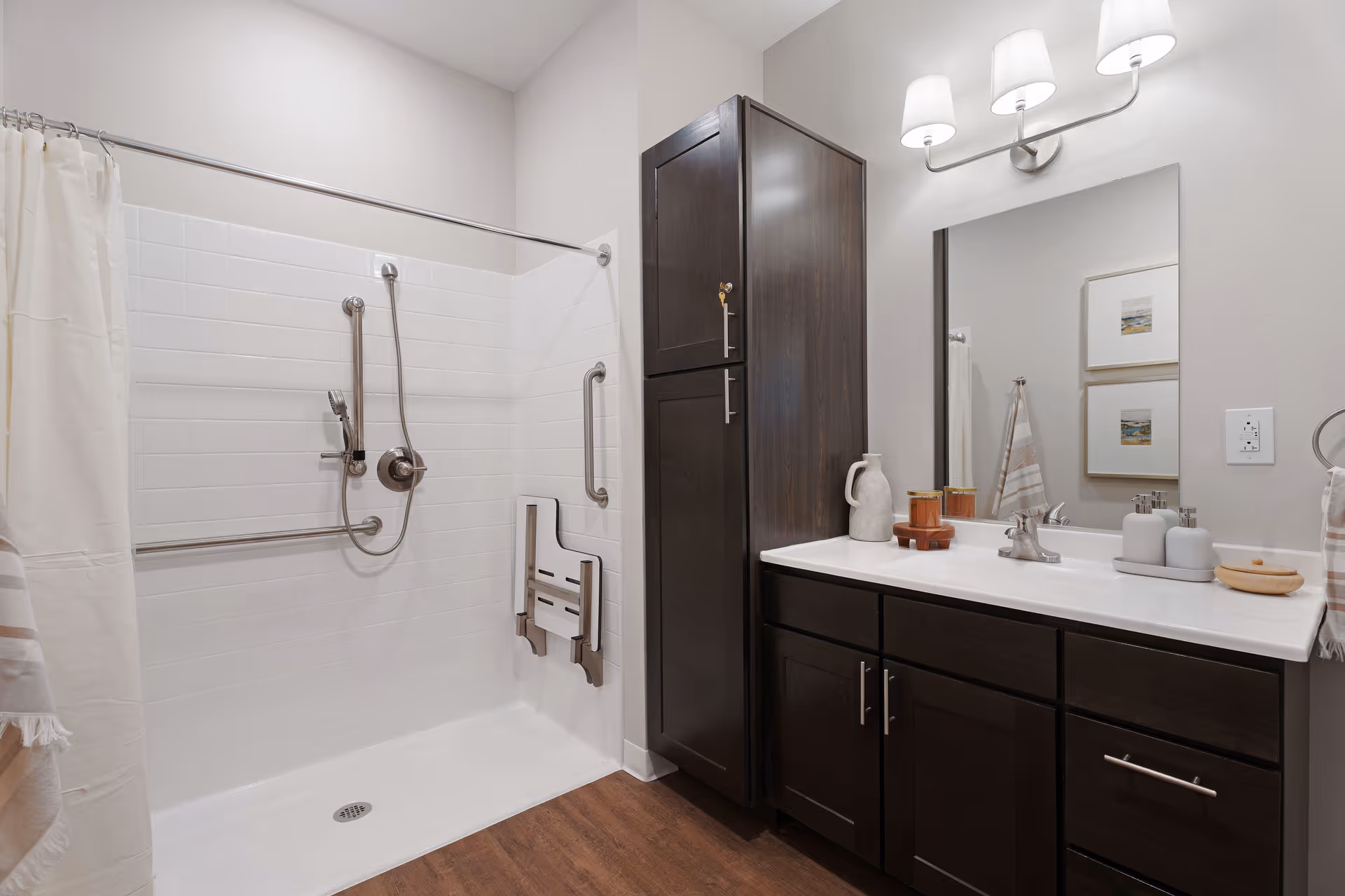 A modern senior living bathroom featuring a walk-in shower with white tiles, grab bars, and a fold-down shower seat. The bathroom has dark wood cabinetry with a white countertop, a large mirror, and wall-mounted light fixtures. The floor is wood-style, and there are decorative items on the countertop and framed pictures on the wall.