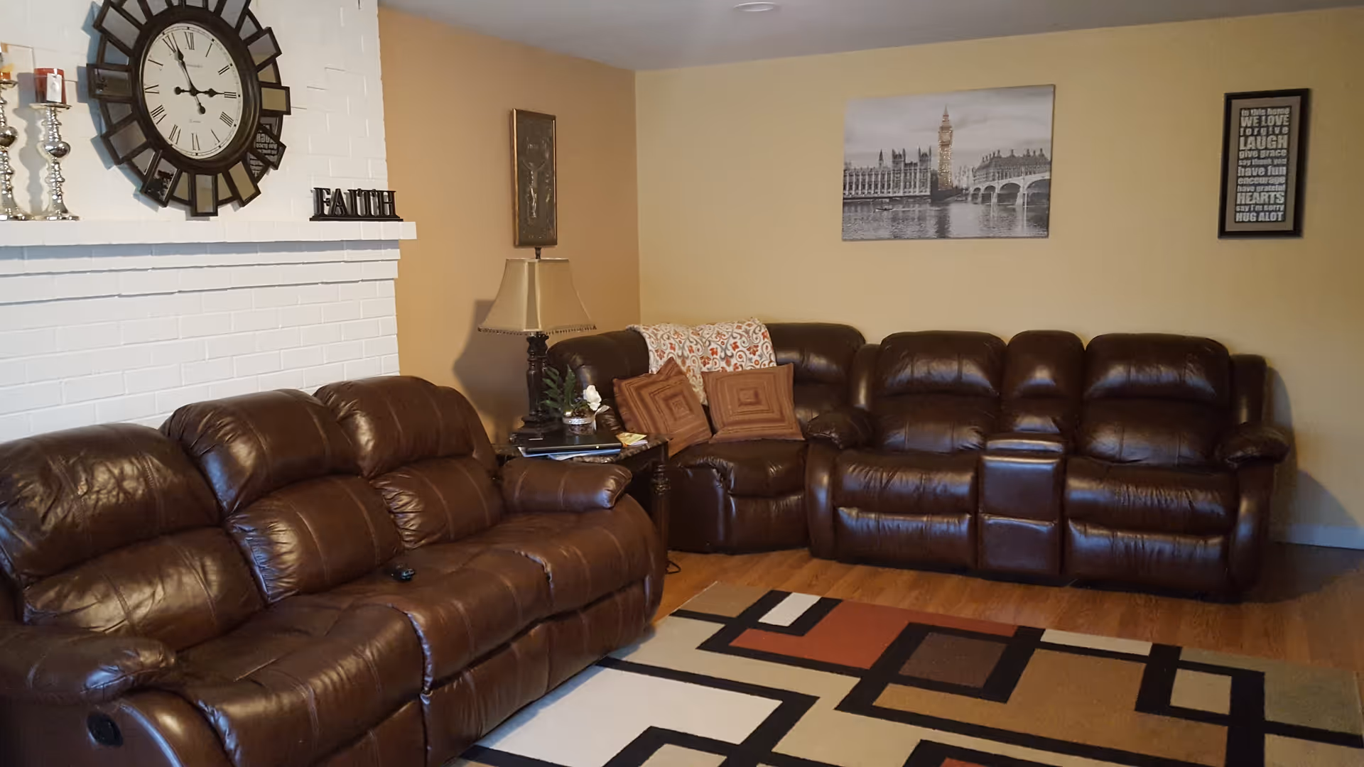 A cozy living room with two brown leather sofas, a patterned area rug, a side table with a lamp and decorative items, a large wall clock above a white brick fireplace, and framed artwork on beige walls.