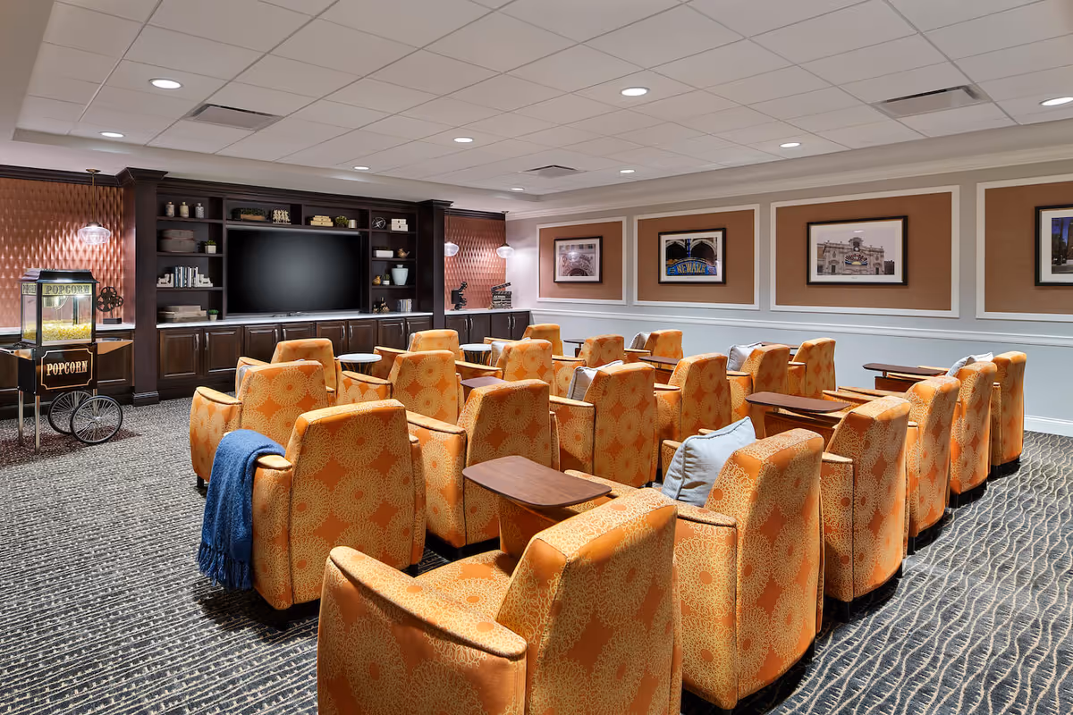 A cozy media room with multiple rows of orange patterned armchairs facing a large flat-screen TV mounted on a dark wooden entertainment center. There is a popcorn machine on the left side of the room, and framed pictures hang on the beige and white walls. The room has a carpeted floor and recessed ceiling lights.